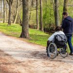 View from back to a woman in wheelchair and a man walking and checking his phone in a park near river at spring