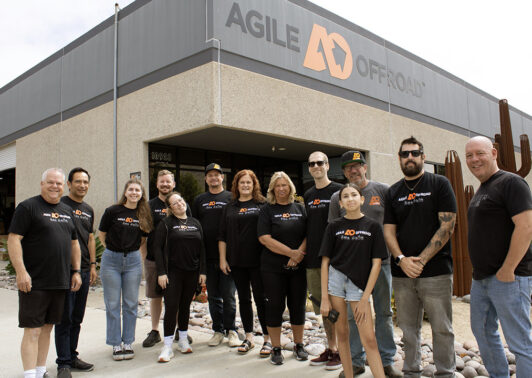 Gregg and his daughter, Kailey, pose alongside the Agile Offroad team, everyone smiling and wearing matching black company T-shirts. They stand together outside the Agile Offroad warehouse beneath a prominent orange-and-black logo sign, accented by decorative metal cactus sculptures.