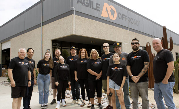 Gregg and his daughter, Kailey, pose alongside the Agile Offroad team, everyone smiling and wearing matching black company T-shirts. They stand together outside the Agile Offroad warehouse beneath a prominent orange-and-black logo sign, accented by decorative metal cactus sculptures.