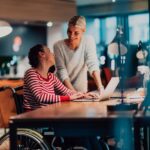 A woman in a wheelchair is working on a laptop, smiling as she looks toward her colleague.