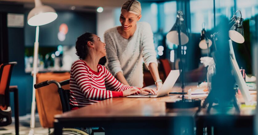 A woman in a wheelchair is working on a laptop, smiling as she looks toward her colleague.