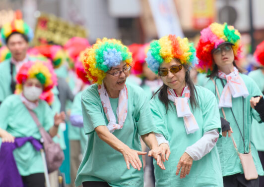 A DeafBlind woman holding hands with her guide, wearing clourful wigs as they dance together during a Kagoshima Ohara Festival.