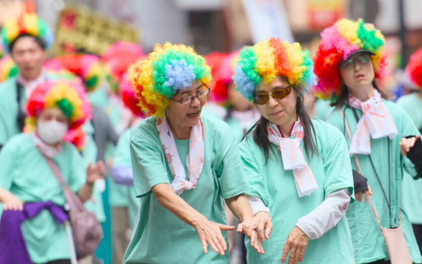A DeafBlind woman holding hands with her guide, wearing clourful wigs as they dance together during a Kagoshima Ohara Festival.
