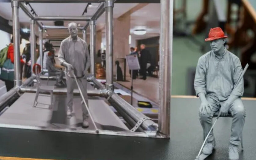 A model of a man with a red had sat on a chair with a white cane next to a photo of a similar figure