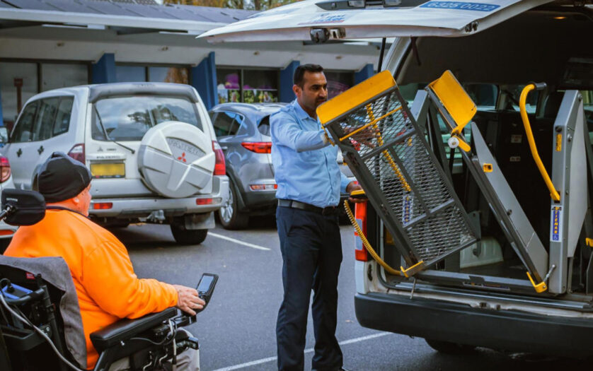 A wheelchair user waits beside a taxi while the driver pulls out the wheelchair lift.