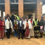 A group of adults stand together outside a brick building, some wearing reflective vests and others in patterned clothing, posing for a group photo