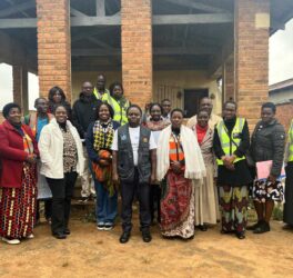 A group of adults stand together outside a brick building, some wearing reflective vests and others in patterned clothing, posing for a group photo