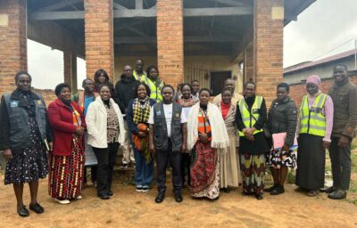 A group of adults stand together outside a brick building, some wearing reflective vests and others in patterned clothing, posing for a group photo