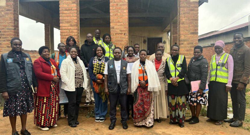A group of adults stand together outside a brick building, some wearing reflective vests and others in patterned clothing, posing for a group photo