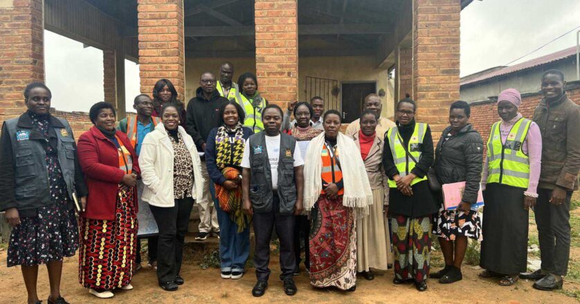 A group of adults stand together outside a brick building, some wearing reflective vests and others in patterned clothing, posing for a group photo