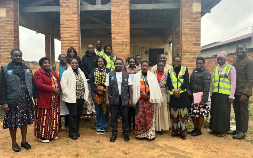 A group of adults stand together outside a brick building, some wearing reflective vests and others in patterned clothing, posing for a group photo