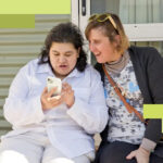 A young woman shows her friend what she's doing online as they sit together on a porch
