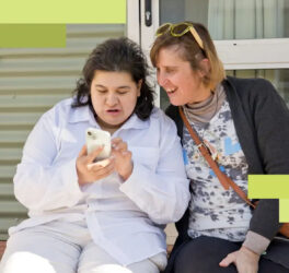 A young woman shows her friend what she's doing online as they sit together on a porch