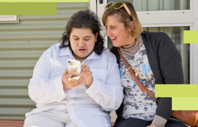 A young woman shows her friend what she's doing online as they sit together on a porch