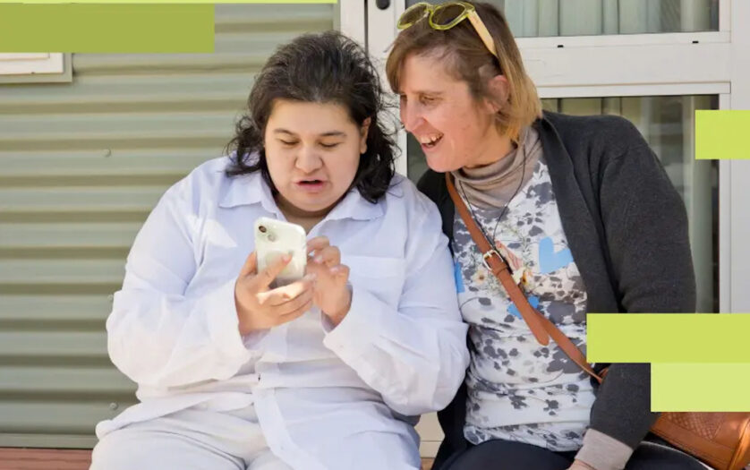 A young woman shows her friend what she's doing online as they sit together on a porch