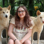 A photo of Grace, a young person with long brown hair and glasses wearing a white flowing top as they sit smiling between two light-coloured dogs.