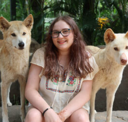 A photo of Grace, a young person with long brown hair and glasses wearing a white flowing top as they sit smiling between two light-coloured dogs.