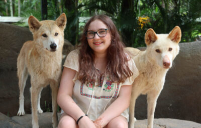 A photo of Grace, a young person with long brown hair and glasses wearing a white flowing top as they sit smiling between two light-coloured dogs.
