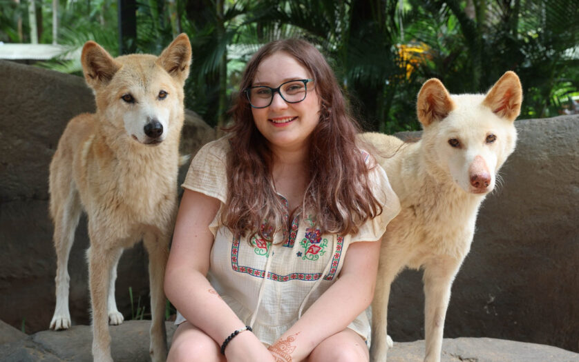 A photo of Grace, a young person with long brown hair and glasses wearing a white flowing top as they sit smiling between two light-coloured dogs.