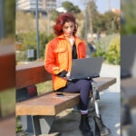 A young girl sits in a park, working on her laptop, surrounded by nature. Her colorful outfit and appearance reflect her energy and optimism. With a prosthetic leg, she proves that disability is no barrier to connecting with nature and embracing digitalization. This photo captures the harmony of technology, youth, and the outdoors—a powerful image of resilience and progress.