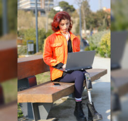 A young girl sits in a park, working on her laptop, surrounded by nature. Her colorful outfit and appearance reflect her energy and optimism. With a prosthetic leg, she proves that disability is no barrier to connecting with nature and embracing digitalization. This photo captures the harmony of technology, youth, and the outdoors—a powerful image of resilience and progress.