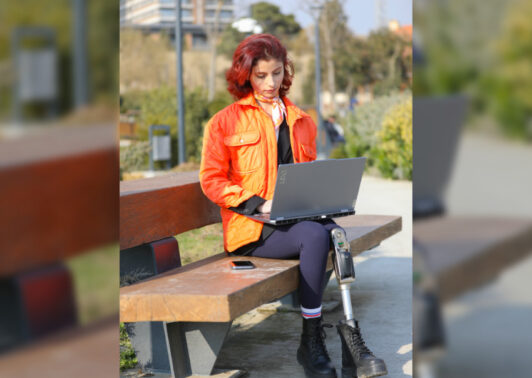 A young girl sits in a park, working on her laptop, surrounded by nature. Her colorful outfit and appearance reflect her energy and optimism. With a prosthetic leg, she proves that disability is no barrier to connecting with nature and embracing digitalization. This photo captures the harmony of technology, youth, and the outdoors—a powerful image of resilience and progress.