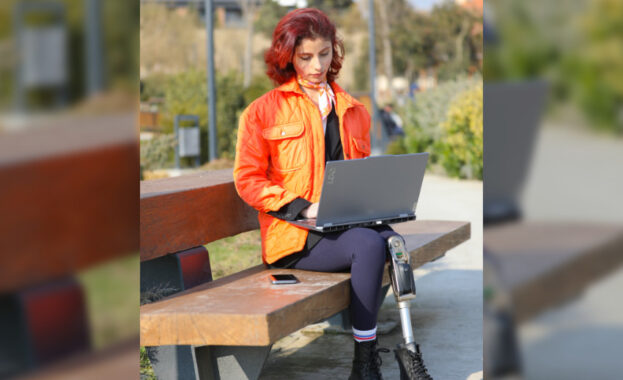 A young girl sits in a park, working on her laptop, surrounded by nature. Her colorful outfit and appearance reflect her energy and optimism. With a prosthetic leg, she proves that disability is no barrier to connecting with nature and embracing digitalization. This photo captures the harmony of technology, youth, and the outdoors—a powerful image of resilience and progress.