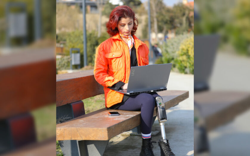 A young girl sits in a park, working on her laptop, surrounded by nature. Her colorful outfit and appearance reflect her energy and optimism. With a prosthetic leg, she proves that disability is no barrier to connecting with nature and embracing digitalization. This photo captures the harmony of technology, youth, and the outdoors—a powerful image of resilience and progress.