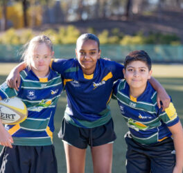 Three children wearing rugby uniforms in a huddle on a playing field facing towards the camera.