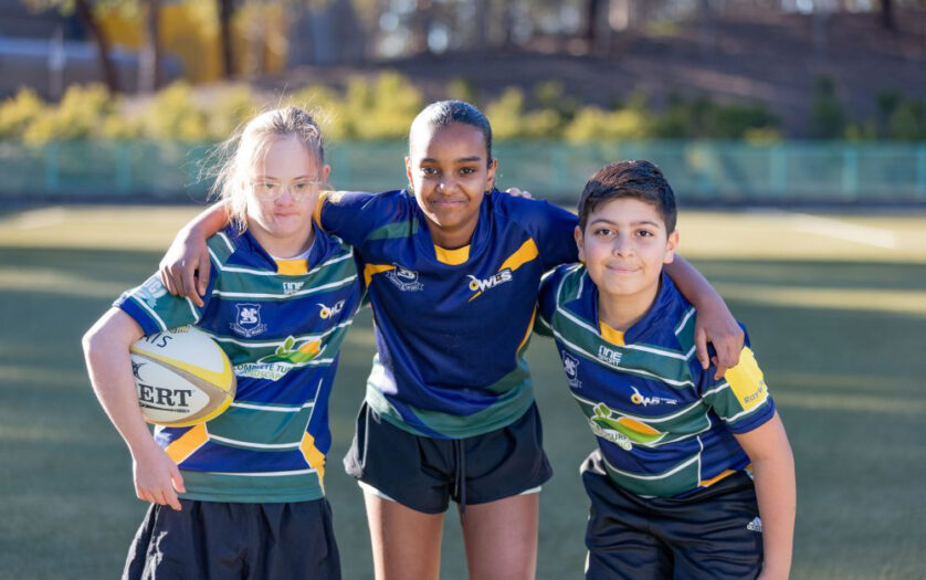 Three children wearing rugby uniforms in a huddle on a playing field facing towards the camera.