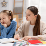 Mother and her upset daughter reading book at table indoors