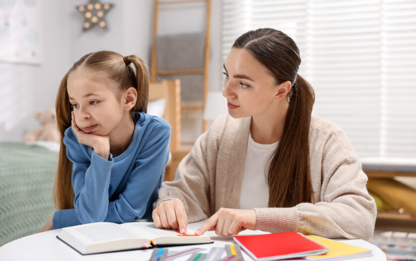 Mother and her upset daughter reading book at table indoors