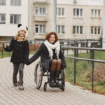 Mother in a wheelchair with her daughter walking outside at park.