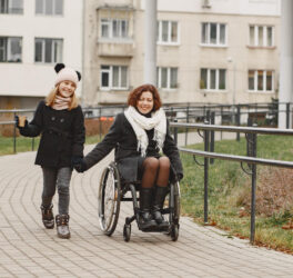 Mother in a wheelchair with her daughter walking outside at park.