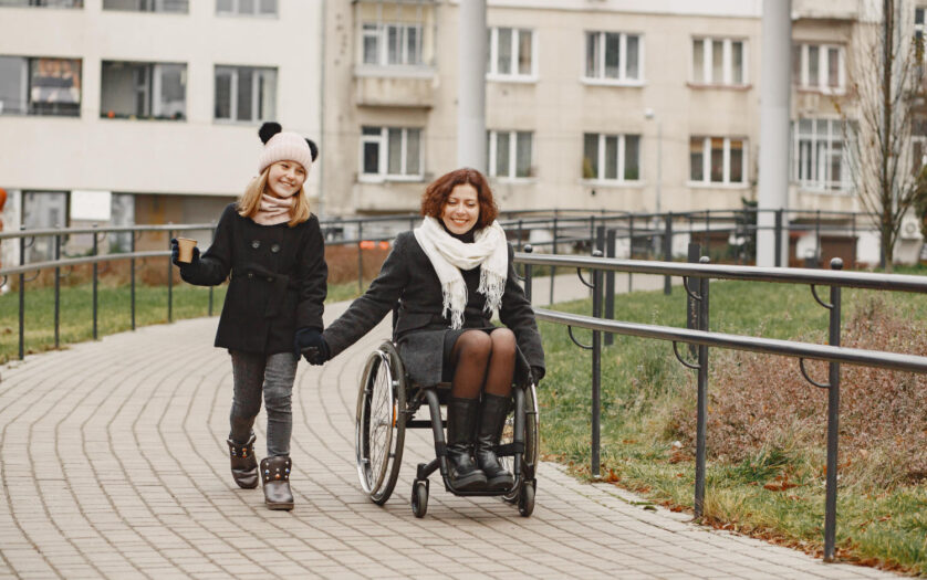 Mother in a wheelchair with her daughter walking outside at park.