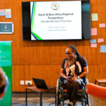 Ms Ogolo-Dickson presenting at an indoor event, with a presentation screen behind them and a lectern and laptop nearby.
