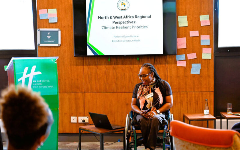 Ms Ogolo-Dickson presenting at an indoor event, with a presentation screen behind them and a lectern and laptop nearby.