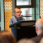 A photo of Jerzy, a 10-year-old boy swith short dark blond hair wearing a blue button-up top and glasses. He is standing at a lectern and speaking into a microphone in front of a crowd