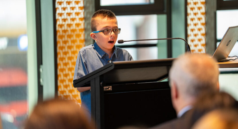 A photo of Jerzy, a 10-year-old boy swith short dark blond hair wearing a blue button-up top and glasses. He is standing at a lectern and speaking into a microphone in front of a crowd