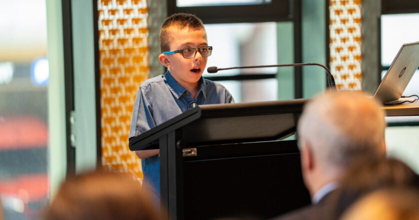 A photo of Jerzy, a 10-year-old boy swith short dark blond hair wearing a blue button-up top and glasses. He is standing at a lectern and speaking into a microphone in front of a crowd