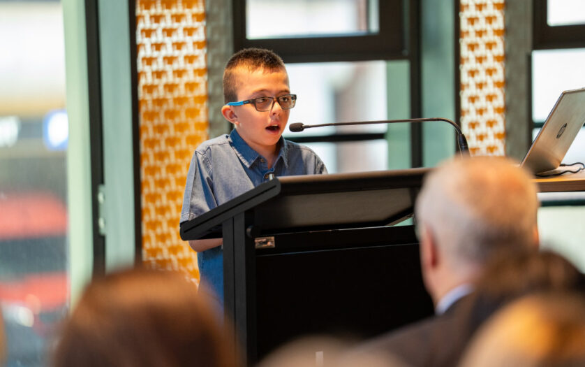 A photo of Jerzy, a 10-year-old boy swith short dark blond hair wearing a blue button-up top and glasses. He is standing at a lectern and speaking into a microphone in front of a crowd