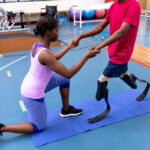 Side view of female physiotherapist helping disabled man walk with prosthetic leg in sports center.