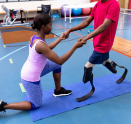 Side view of female physiotherapist helping disabled man walk with prosthetic leg in sports center.