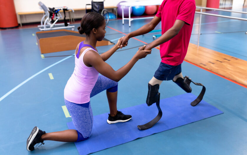 Side view of female physiotherapist helping disabled man walk with prosthetic leg in sports center.