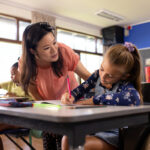 Diverse female teacher and schoolgirl in wheelchair in elementary school class.
