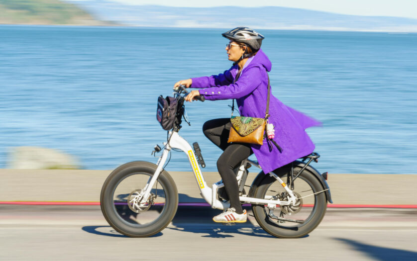 A woman rides an electric bicycle along the waterfront, wearing a purple coat and helmet, on a sunny day.