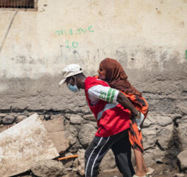 A person in a red vest and face mask carries another person on their back along a rough, debris-strewn street next to a worn wall.