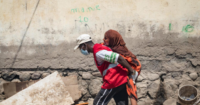 A person in a red vest and face mask carries another person on their back along a rough, debris-strewn street next to a worn wall.