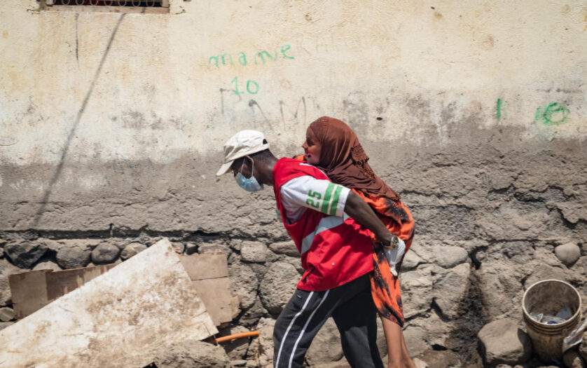 A person in a red vest and face mask carries another person on their back along a rough, debris-strewn street next to a worn wall.